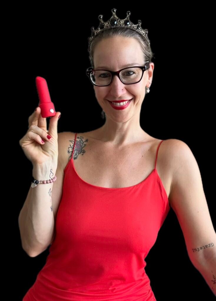 Pleasure Toy Queen Patricia holding a red lipstick vibrator wearing a red dress and her crown.